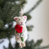 A wool felt sheep ornament holding a heart hangs on a cheerful Christmas tree. This soft, fluffy ornament features tiny pink ears, a pink stitched nose, and closed stitched eyes, with a jute string for hanging.