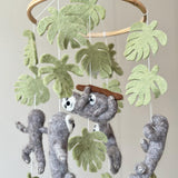 A Wool Felt Sloth Baby Mobile is presented against a light, neutral background. It features a wooden frame from which several gray wool felt sloths are suspended by white cotton threads. One sloth hangs horizontally, while other sloths hang vertically with limbs wrapped in flexible wire, allowing them to be bent and twisted. The sloths have visible white and black detailing on their faces.