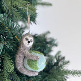 A handcrafted felt sloth ornament is showcased on a lush green Christmas tree. This unique Christmas ornament features a gray sloth with a light gray face, closed eyes, and a smiling mouth, gently embracing a blue and green globe. It evokes a whimsical sense of environmental awareness, perfect as an Earth-holding decor piece.