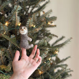 A hand holds a small, felt sloth finger puppet against the backdrop of a Christmas tree. The needle felted sloth is depicted in profile view, facing right, with a brown body and lighter-colored face featuring two small eyes and a small smile. It's made from wool felt, creating a soft and cozy atmosphere.