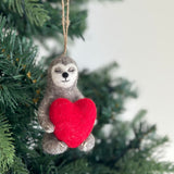A heartwarming wool felt sloth holiday heart ornament hangs on a Christmas tree, featuring a gray and white sloth. This handcrafted sloth holds a vibrant red heart. Its soft wool felt texture and attached jute string are clearly visible against the evergreen needles.