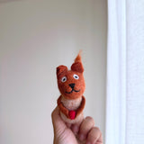 A handmade Needle Felted Squirrel with Acorn finger puppet is held up by a female hand against a bright white wall and sheer curtain, enhanced by natural light. This unique squirrel puppet, crafted in Nepal, is light brown with a darker head and tail, featuring small ears, a stitched smile, and holding a red acorn in its paws. Its curious and playful expression makes it a charming felt toy for kids.