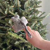 A handcrafted wool felt sugar glider finger puppet is showcased by a female hand, set against a blurred background of a Christmas tree adorned with warm, festive lights. This playful 3D needle felted sugar glider features a gray body with a dark gray stripe down its back and delicate pink details, highlighting its intricate design.