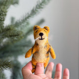 A female hand holds a charming, three-dimensional needle felted tiger finger puppet against a blurred Christmas tree backdrop. This handmade wool felt tiger finger puppet, skillfully crafted by artisans in Nepal, features a tan body with black stripes, rounded ears, and white whiskers extending from its smiling face with closed eyes.