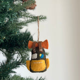 A wool felt travel backpack ornament hangs on a Christmas tree, evoking a sense of wanderlust. The black backpack features a yellow pocket, a light gray square, a yellow and blue circle, and two small white lines. It has a jute string attached for easy hanging.