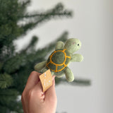 A handcrafted wool felt turtle finger puppet is presented, held by a female hand, with a blurred Christmas tree in the background. This unique felt turtle finger puppet is light green with a darker green shell outlined in yellow. It features small, rounded flippers and a sweet expression, reflecting its playful and artisan-made design.