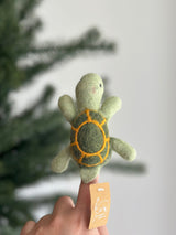A whimsical, handcrafted wool felt turtle finger puppet, perfect for imaginative finger play, is held upright on a finger of a female hand. This unique felt turtle features a light-green head, body, and flippers, complemented by a dark green shell with orange line-drawn segments. A blurred Christmas tree is visible in the background, along with the product's tag.