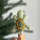 A whimsical, handcrafted wool felt turtle finger puppet, perfect for imaginative finger play, is held upright on a finger of a female hand. This unique felt turtle features a light-green head, body, and flippers, complemented by a dark green shell with orange line-drawn segments. A blurred Christmas tree is visible in the background, along with the product's tag.