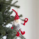 A handmade wool felt Pickleball Gnome Ornament hangs from a jute string on a Christmas tree. This unique Christmas ornament has a tall, red polka-dotted hat, a fluffy white beard, a gray body with red arms, and candy cane-striped legs.