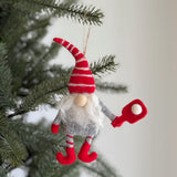 A playful wool felt pickleball gnome ornament hangs from a pine branch, adding festive warmth to a blurred Christmas tree. This unique Christmas ornament features a gnome in a gray suit with red cuffs and shoes, a tall, floppy red hat with white stripes, and a fluffy white beard. It holds a red pickleball paddle with a white felt ball.