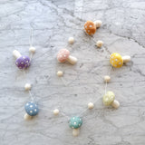A Felt Pastel Mushroom Garland rests on a white marble surface. It features seven handcrafted wool felt mushrooms in pastel colors like blush, honey, mint, lilac, and blue. Each mushroom has a rounded cap with white spots and a cylindrical stem, interspersed with small ivory felt balls.