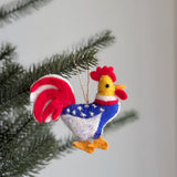 A handmade wool felt patriotic chicken ornament hangs from a pine tree branch, set against a blurred white background. This unique Christmas ornament features a red crest, yellow beak, and neck. Its body is blue with white stars, complemented by white legs, yellow feet, and red and white striped tail feathers, with a jute string for hanging.