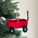 A red wool felt wagon ornament hangs playfully from the green branches of a Christmas tree. This unique, handcrafted ornament features a bright red body, black wheels with white centers, and a gray handle, with a rustic twine loop for hanging. It is made through fair-trade, eco-friendly practices.