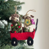 A bright red wool felt wagon ornament, handcrafted by fair-trade artisans, hangs on a green Christmas tree branch. Inside, two charming wool felt sheep finger puppets are nestled: a light brown sheep holding a green felt garland with red berries, and a white sheep clutching a bouquet of colorful felt flowers. The wagon features black wheels and a grey handle, adding a playful touch to the holiday decor.