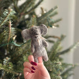 A light gray wool felt elephant finger puppet, handcrafted from textured wool, is held by a female hand with red polished nails. Its distinctive ears and tusks are visible against a blurred Christmas tree background adorned with lights, creating a cozy and festive scene.
