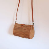 The Bali Envelope Rattan Crossbody Bag is hang from top against a white wall, showcasing its natural rattan Ata grass texture and real leather straps. The bag's bohemian design is enhanced by a unique Batik lining. Made from eco-friendly materials, this handcrafted piece is perfect for any occasion. With a sturdy base and detachable strap, it can stand on its own or be carried as a clutch.