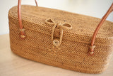 A woven, light brown wicker bag rests on a wooden surface, featuring a handle made from the same material as the body secured with a leather snap closure. A visible bow-tie detail adds decorative appeal. The bag's shape is petite and relaxed, with a smooth rattan finish that won't catch on clothing.