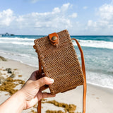A person holding a woven rattan crossbody cell phone bag against a beach backdrop. The brown bag features a visible snap closure, indicating it's secure and adjustable, with a leather strap for comfort and versatility. The sandy beach background evokes a relaxed atmosphere.
