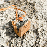 A small, handcrafted rattan cellphone bag rests on a rocky surface, showcasing its rectangular shape and distinctive batik pattern. The bag features a snap closure and adjustable strap, made from high-quality materials that evoke a sense of nature and traditional craftsmanship.
