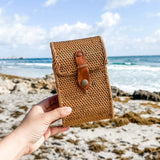 A model holds the Bali Rattan Crossbody Cellphone Bag - Ella, made of natural rattan material with a leather strap and snap closure. The bag features a smooth finish and is lined with Batik cotton for timeless style and durability.