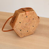 A woven rattan crossbody bag, Bali Rattan Crossbody Bag - Hexagon, rests on a wooden surface. The hexagonal-shaped bag features a unique circular pattern at its center and is adorned with an adjustable brown leather strap.