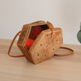 A woven rattan crossbody bag with a hexagonal shape is placed on a wooden surface against a white background. The bag features a red batik lining visible through the top, an adjustable brown leather strap made of natural materials, and a secure snap closure.