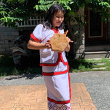 A woman stands on a sidewalk next to a house, wearing a traditional Bali dress and holding a woven rattan crossbody bag with a hexagonal shape. The bag features an adjustable leather strap made from natural materials. The background shows a residential area with trees and a building, suggesting an everyday setting.