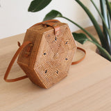 A woven rattan crossbody bag sits on a wooden surface, showcasing its hexagonal shape and batik lining visible through the weave. The adjustable brown leather strap adds versatility for carrying options. Handcrafted with an artisan's imprint, this Bali-made bag exudes a casual, bohemian vibe.