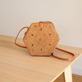 A woven rattan crossbody bag sits on a wooden surface, showcasing its hexagonal shape and unique design element featuring a circular pattern at the center of each side. The bag is adorned with an adjustable brown leather strap, allowing for different carrying styles. The background is minimalistic, with a plain wall and a portion of a table or desk, suggesting a clean and organized setting.