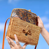 A woven crossbody bag, crafted from rattan or similar material, is held by a person against a blue sky background. The bag features an adjustable leather strap with a detachable shoulder strap, showcasing its floral pattern and textured design.