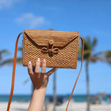 A person holding up a woven crossbody bag against a beach backdrop, featuring a rattan or straw material with a textured weave pattern. The bag has an adjustable leather strap with a metal buckle closure and a small decorative element on the front.