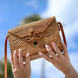 A person holding a crossbody bag made from natural materials, resembling rattan or a similar texture. The bag has an adjustable leather strap and measures approximately 8.6 inches by 4.75 inches by 6.25 inches (22 cm x 8 cm x 14.5 cm). It features a simple design with rounded corners and a decorative bow at one corner, showcasing visible texture on its surface.