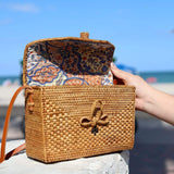 A person holding an open woven crossbody bag, crafted from natural materials like rattan or wicker, featuring a floral pattern. The adjustable leather strap has a 22-inch drop, with the bag measuring approximately 8.6 inches in height and 4.75 inches in width. Set against a casual outdoor background with sand and blue sky.