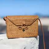 A woven crossbody bag, made from light-colored rattan material with a visible weaving pattern, rests on a surface against a clear sky backdrop suggesting an outdoor beach setting. The rectangular-shaped bag features rounded corners, a flap closure secured by a buckle, and a small leather strap attached to the top edge for adjustable wear. The woven texture and stitching details are prominently displayed, showcasing the natural material's unique characteristics.