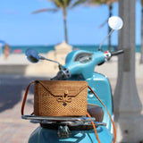 A Bali Rattan Crossbody Bag rests on the seat of a parked motor scooter, positioned in front of a beach setting with palm trees. The bag is woven from natural rattan material, featuring an adjustable leather strap made from real leather. Its compact rectangular shape and earthy tones blend seamlessly into the tropical surroundings.