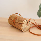 A woven rattan crossbody bag rests on a wooden surface, its cylindrical shape with tapered top and earthy brown color giving it an organic look. The bag features an adjustable leather strap for comfort and a snap closure at the front, showcasing its eco-friendly design.