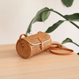 A woven rattan crossbody bag rests on a wooden surface, surrounded by a plant's natural beauty. The Bali Rattan Crossbody Bag - Tapang features a cylindrical shape, brown color, and natural materials. An adjustable strap and snap closure add functionality, while the unique Batik lining inside adds elegance.