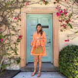 A Bali Rattan Handbag - Capri, handwoven from natural rattan Ata grass in Bali, is held by a woman in a vibrant orange dress. She stands before a famous turquoise door in Palm Beach, Florida, which is framed by climbing pink bougainvillea flowers. The bag is detailed with real leather handles and a lid that has a hook closure.