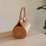 A woven handbag made from rattan, resting on a flat surface against a plain background. The bag has a cylindrical shape with a slightly tapered top, featuring a textured, woven pattern throughout its body and rattan handles. Its interior is lined with a fabric displaying a batik design.