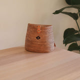 A small, woven rattan backpack rests on a wooden surface, showcasing its rectangular shape with rounded corners. The brown color of the rattan material is visible, along with its textured weave. A decorative emblem adorns the front, and the adjustable leather straps are fully visible. Inside, the unique batik lining adds an extra touch of elegance.