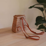 A woven rattan mini backpack rests on a wooden surface, showcasing its rectangular shape and adjustable straps with buckles. The bag features a unique batik lining that adds a touch of color to the neutral material. The background is simple, with a plant visible in the top left corner, suggesting an indoor setting.