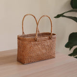 A woven rattan handbag, empty and centered on a table or countertop, features two handles at the top. The background is a simple indoor setting with a plant visible on the left side.