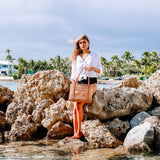 A woman standing on rocks near a body of water, holding a Bali Rattan Woven Tote Bag with leather shoulder straps. The bag appears to be made from natural materials with a woven design. A clear sky and palm trees are visible in the background, suggesting a tropical location.