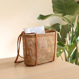 A wicker basket, resembling a tote bag, rests on a wooden surface. The basket features a brown leather strap across the top, suggesting it can be carried over the shoulder. A potted plant is visible in the background, adding a touch of greenery to the scene.