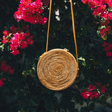 A handmade Bali round rattan crossbody bag, featuring a woven texture, is hanging against a wall of Bougainvillea under daylight. The product boasts vibrant pink Bougainvillea flowers in the background. This eco-friendly and stylish handwoven bag has an adjustable leather strap and distinctive batik lining inside.