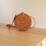 A beautiful Bali Round Rattan Crossbody Bag rests on a wooden surface, its round shape and brown color showcasing the natural rattan material. The bag features adjustable leather straps at the top, a secure snap closure, and a distinctive Batik lining. Made from eco-friendly materials and handwoven by skilled artisans in Bali, this stunning crossbody bag is perfect for adding a chic bohemian flair to any outfit.