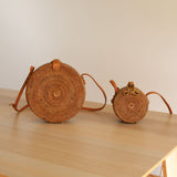 A pair of woven rattan baskets on a light-colored wooden background, each featuring a rustic appearance with natural materials. The larger crossbody bag has a round shape with a flat base and is positioned vertically, while the smaller one sits upright next to it with an adjustable leather strap for wearing on the shoulder. Both have a patterned lining visible through the weave.
