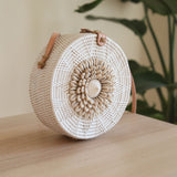 A woven rattan crossbody bag, Bali Seashell Rattan Crossbody Bag - Ava, rests on a table with a plant and white wall background. The bag features seashells in its unique design, has a round shape with a handle for carrying over the shoulder, and is made of natural materials like rattan or bamboo.