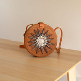 A small, round woven bag with a star-like design on its front rests on a wooden surface. The bag is made from rattan or a similar natural material and features an adjustable strap for crossbody wear. Its batik lining adds a touch of tradition, while the snap closure ensures security.