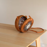 A small, woven purse with a round top rests on a wooden surface, featuring a circular design adorned with seashells. The adjustable strap and snap closure are visible, showcasing the product's eco-friendly and fair-trade craftsmanship.